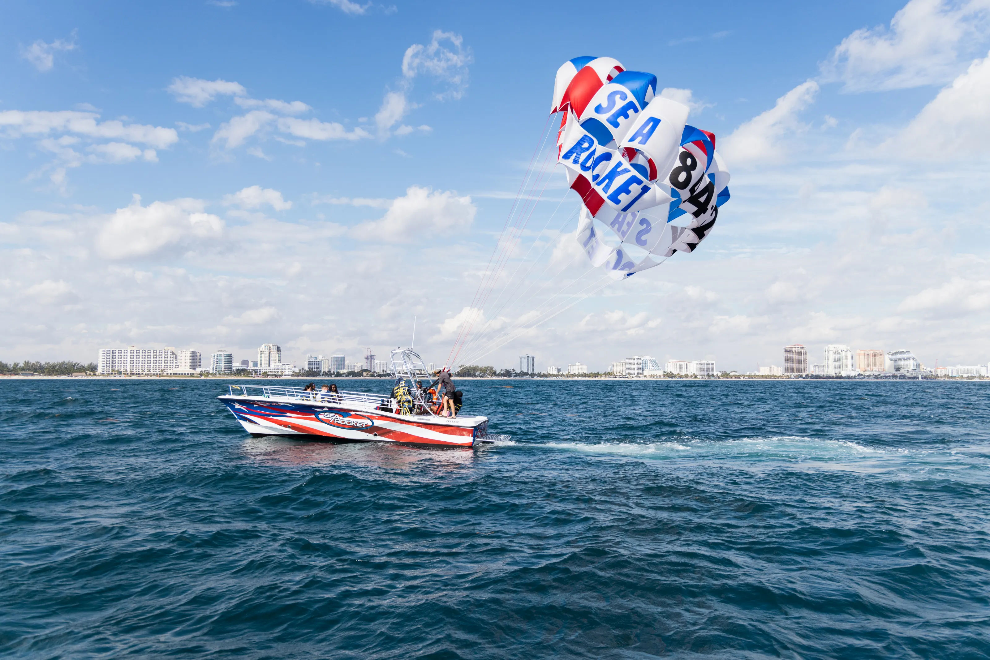 Boat with parasail over ocean, city skyline in background, blue sky with clouds.