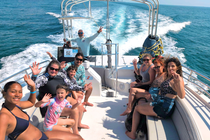 Group of people on a boat in the ocean, smiling and waving at the camera with a clear blue sky.