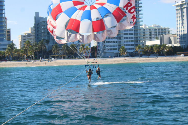 Two people parasailing near a beach with buildings and palm trees in the background.