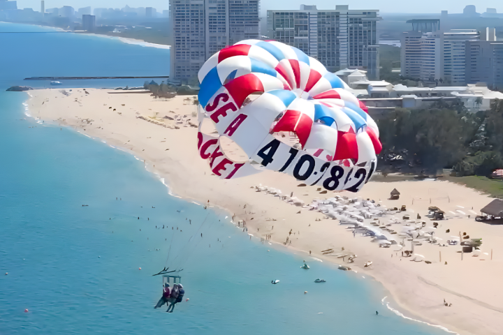 Two people parasailing over a beach with buildings in the background.