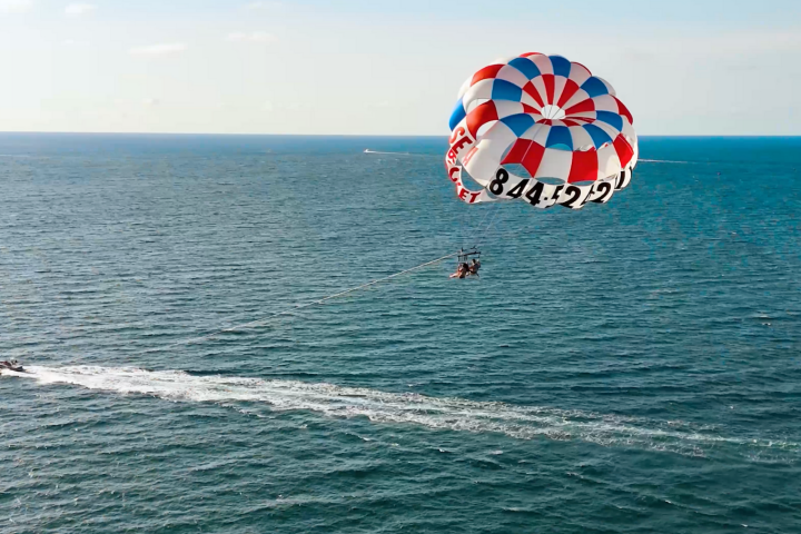 Parasailing over ocean with red, white, blue parachute and speedboat below.