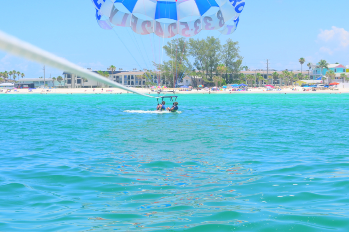 Parasailing over turquoise water with people on a sandy beach in the background.