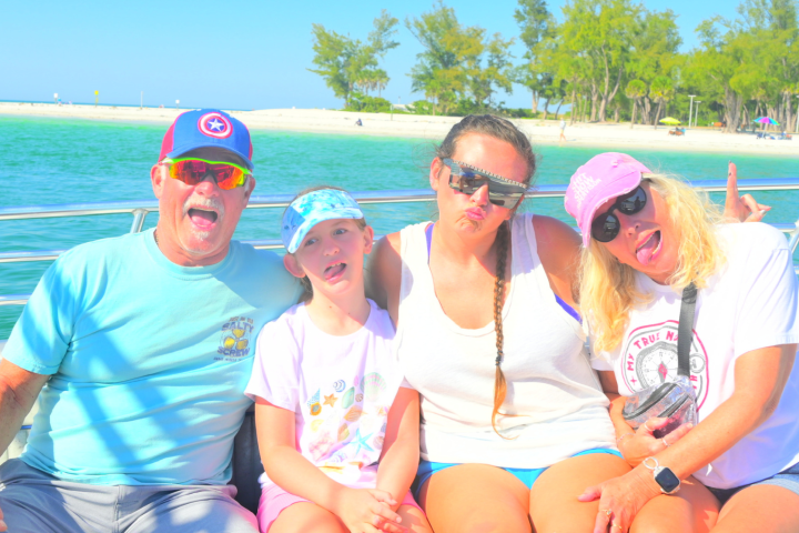 Four people posing and making faces on a boat with a beach and trees in the background.