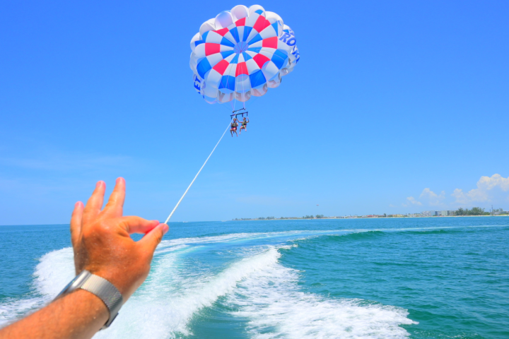 Person paragliding over ocean with hand making OK gesture in foreground.