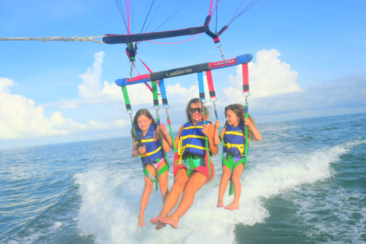 Three people parasailing over the ocean with bright vests and harnesses against a clear, sunny sky.