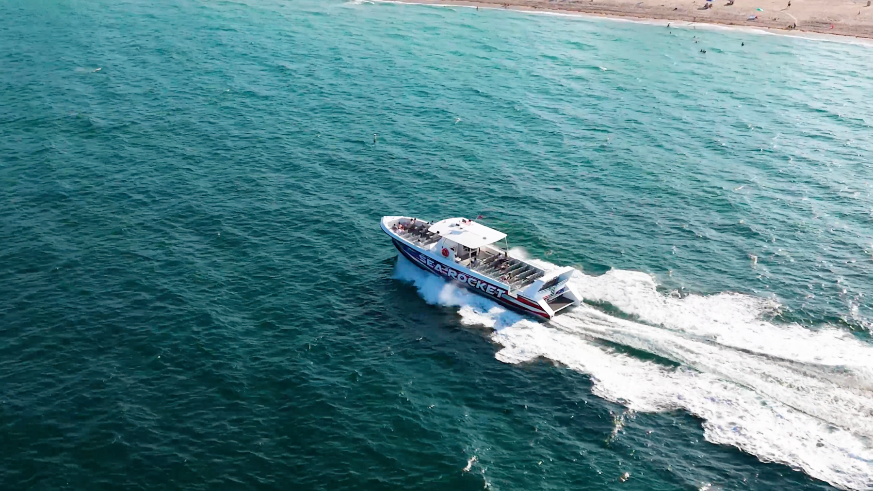 Aerial view of a motorboat speeding across blue ocean waters near a sandy coastline.