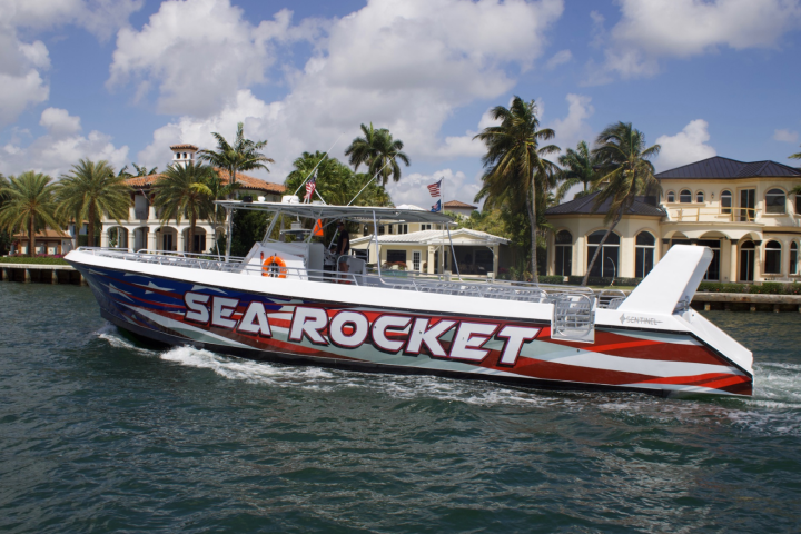 Boat named Sea Rocket on water, palm trees and mansion in background.