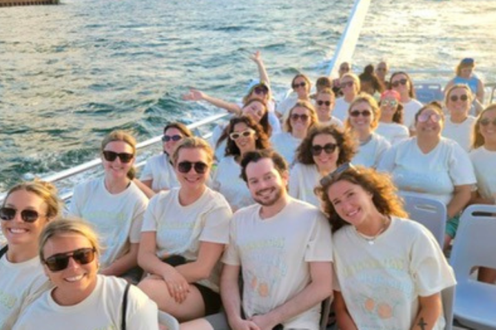 Group of people on a boat, smiling and wearing light-colored shirts, with water in the background.
