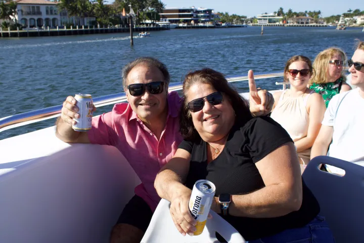 People on a boat enjoying drinks, with a waterfront view and palm trees in the background.