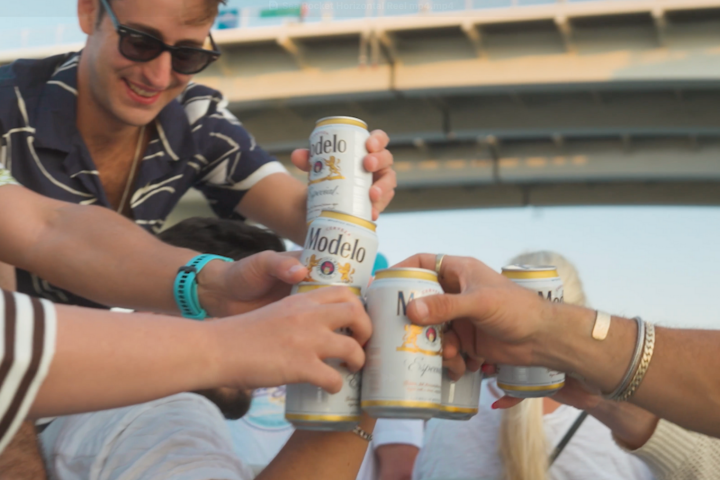 Group of people clinking cans of Modelo beer under a bridge.
