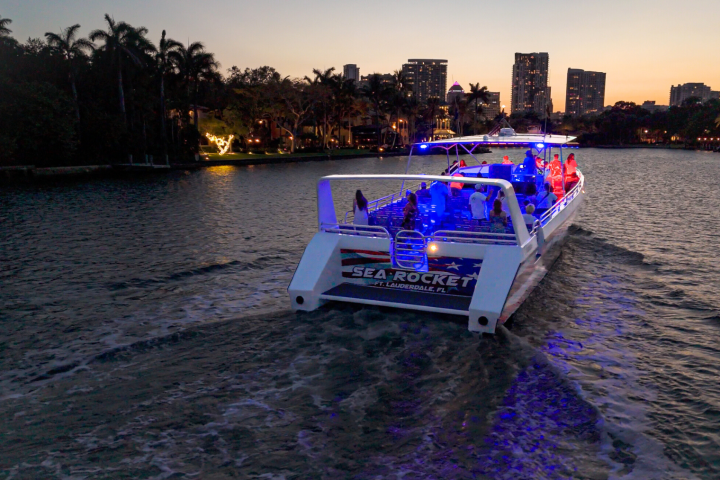Boat with blue lights cruising a river at dusk, city skyline in background.