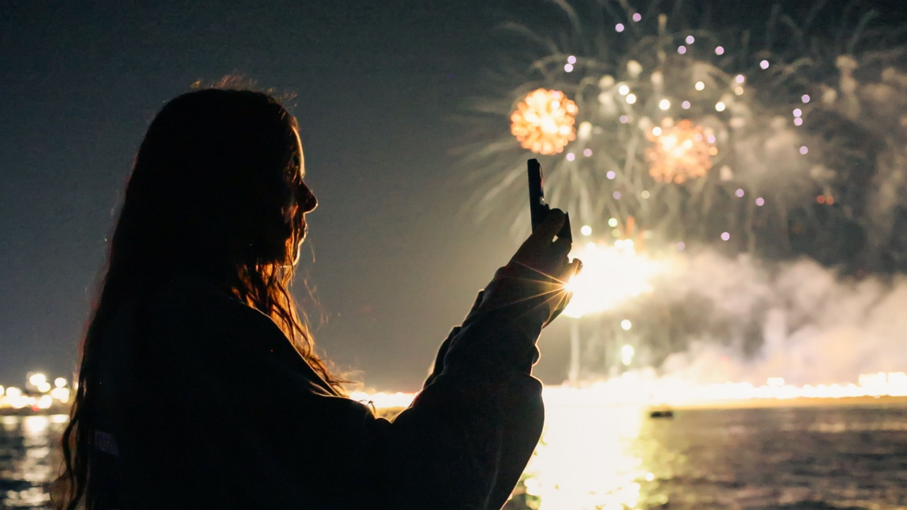 Silhouette of person taking photo of fireworks at night over water.