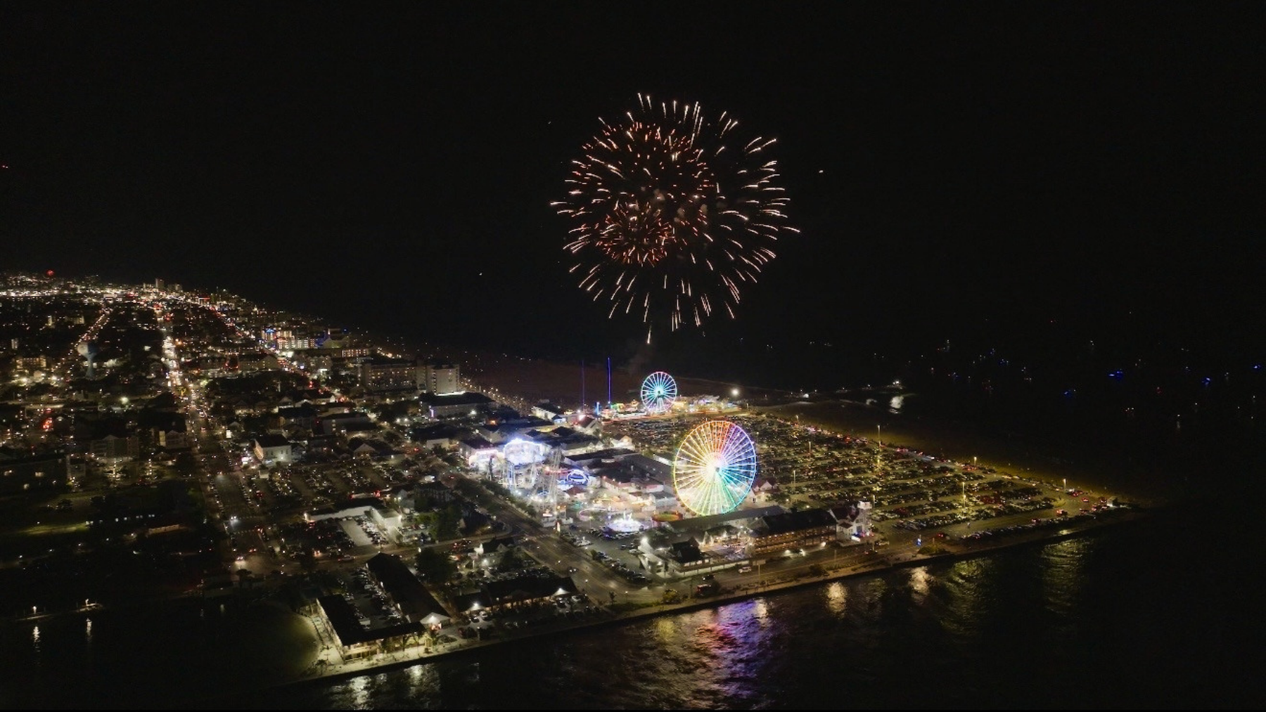 Night aerial view of amusement park with lit Ferris wheel and fireworks above.