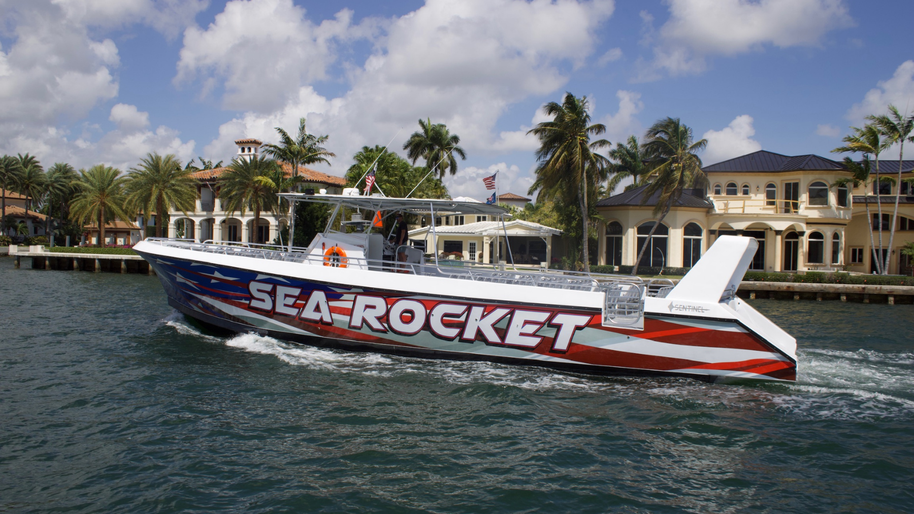 Speedboat with 'Sea Rocket' design on water, palm trees and luxury houses in background.