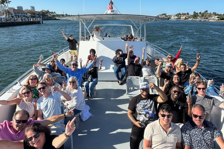 Group of people enjoying a sunny boat ride, smiling and raising hands.