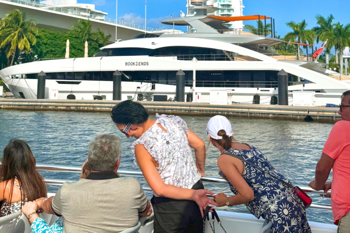 People on a boat observe a large yacht docked near palm trees and greenery.