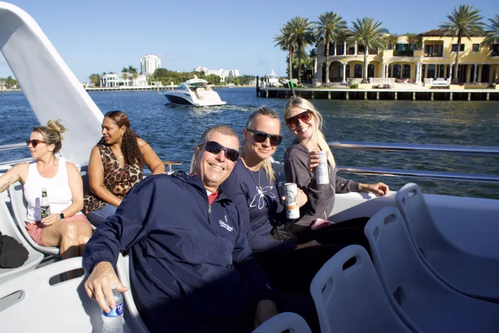 Group of people enjoying a boat ride on a sunny day with a cityscape and palm trees in the background.