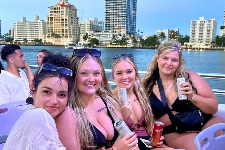 Four women smiling on a boat with cityscape in the background.