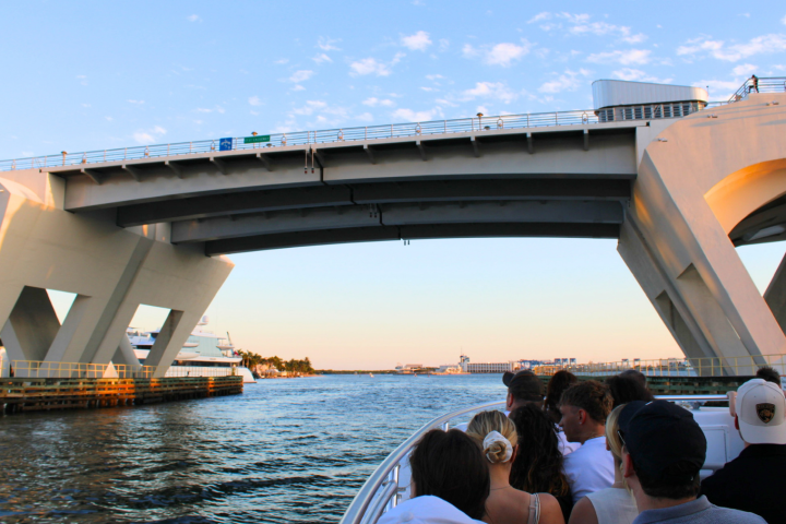 People on a boat passing under a large concrete bridge over a waterway.