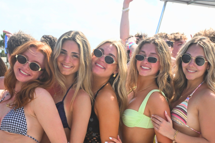 Five smiling women in swimsuits and sunglasses posing in a group.