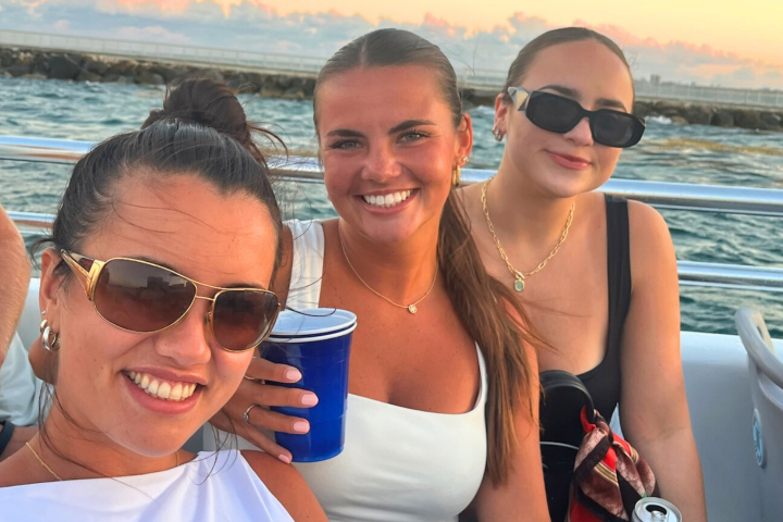 Three women smiling on a boat with water and sunset in the background.
